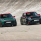 Two all-new MINI John Cooper Works models—an Aceman in Rebel Green and a 3-door Electric in Midnight Black—driving side by side on a steeply banked concrete track.