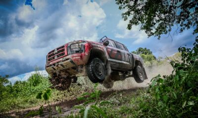 A red Mitsubishi Triton rally truck airborne on a muddy trail, mid-jump during off-road testing, with dirt flying under its tires and dense greenery surrounding the scene.