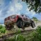 A red Mitsubishi Triton rally truck airborne on a muddy trail, mid-jump during off-road testing, with dirt flying under its tires and dense greenery surrounding the scene.