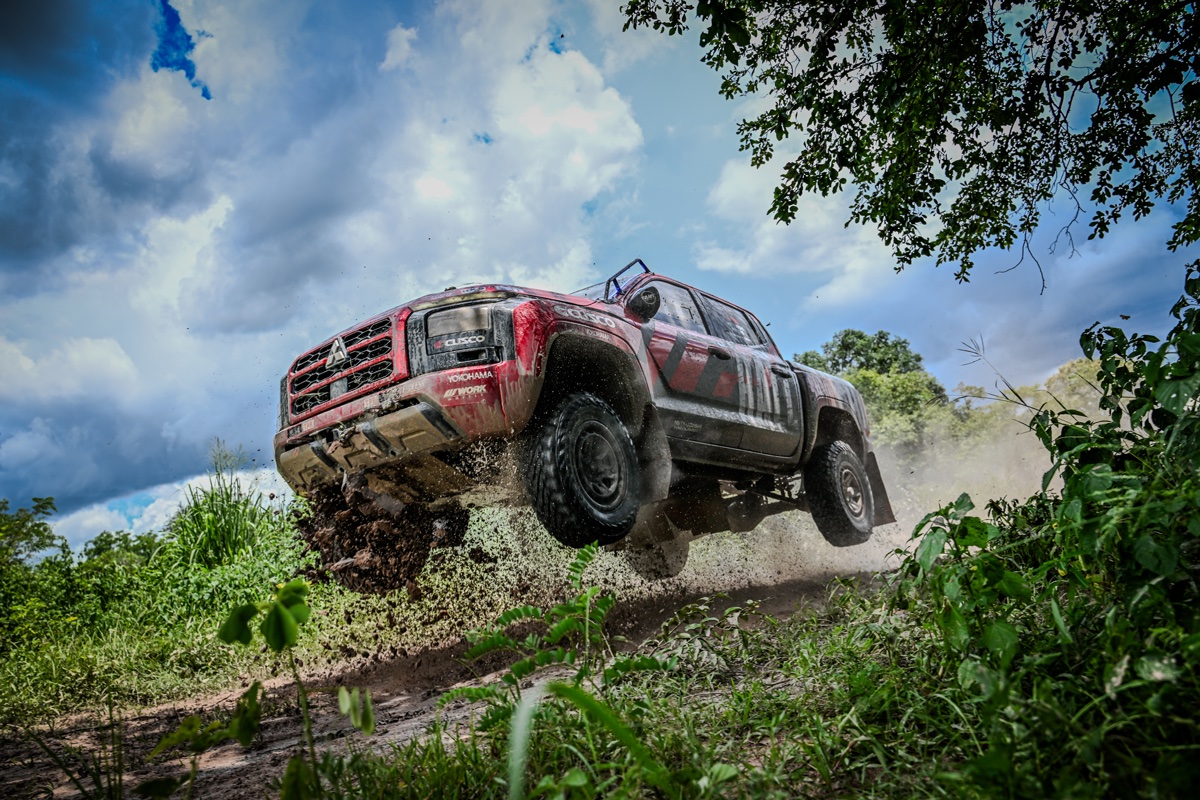 A red Mitsubishi Triton rally truck airborne on a muddy trail, mid-jump during off-road testing, with dirt flying under its tires and dense greenery surrounding the scene.