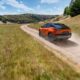 2026 Subaru Uncharted in orange driving on a dusty off-road trail through grassy hills under a bright blue sky.