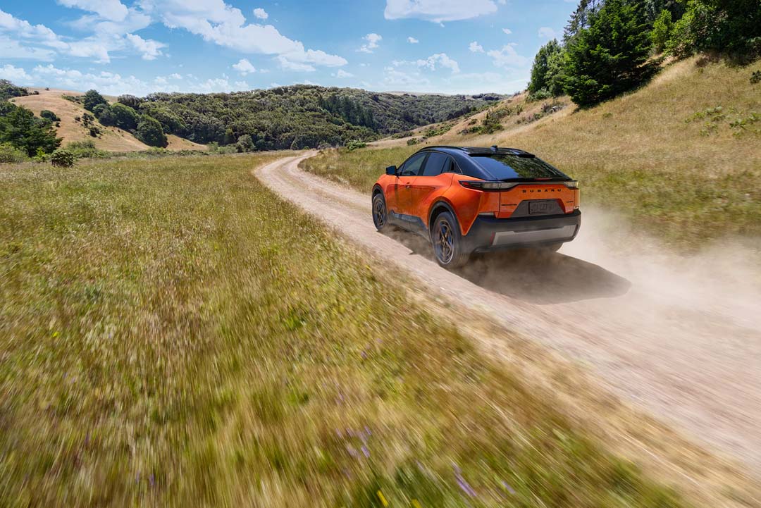 2026 Subaru Uncharted in orange driving on a dusty off-road trail through grassy hills under a bright blue sky.