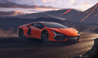 Bright orange Lamborghini Revuelto hybrid supercar parked on a rugged volcanic landscape at sunset, with glowing lava trails on mountains in the background.