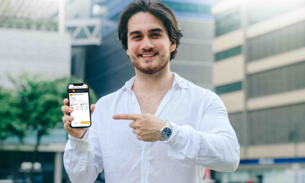 Man in white shirt smiles and points to a smartphone showing the Shell Recharge station on the Evro app.