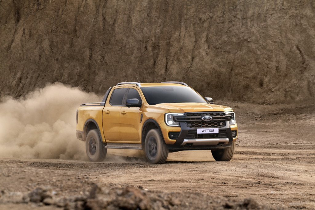 A bold yellow Ford Ranger Wildtrak kicks up dust as it powers through a rugged, dirt-covered trail with rocky terrain and a steep cliffside in the background.