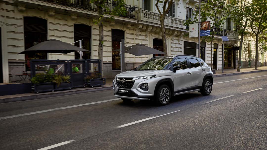 Silver Suzuki Fronx GLX compact SUV driving along a city street lined with outdoor cafes and historic buildings