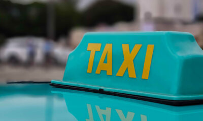 Close-up of a turquoise electric taxi roof sign with yellow "TAXI" letters, showing a clear reflection on the car's glossy surface.