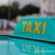 Close-up of a turquoise electric taxi roof sign with yellow "TAXI" letters, showing a clear reflection on the car's glossy surface.
