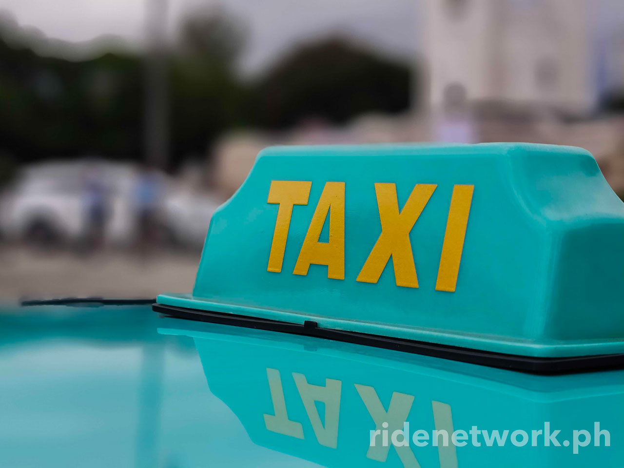 Close-up of a turquoise electric taxi roof sign with yellow "TAXI" letters, showing a clear reflection on the car's glossy surface.