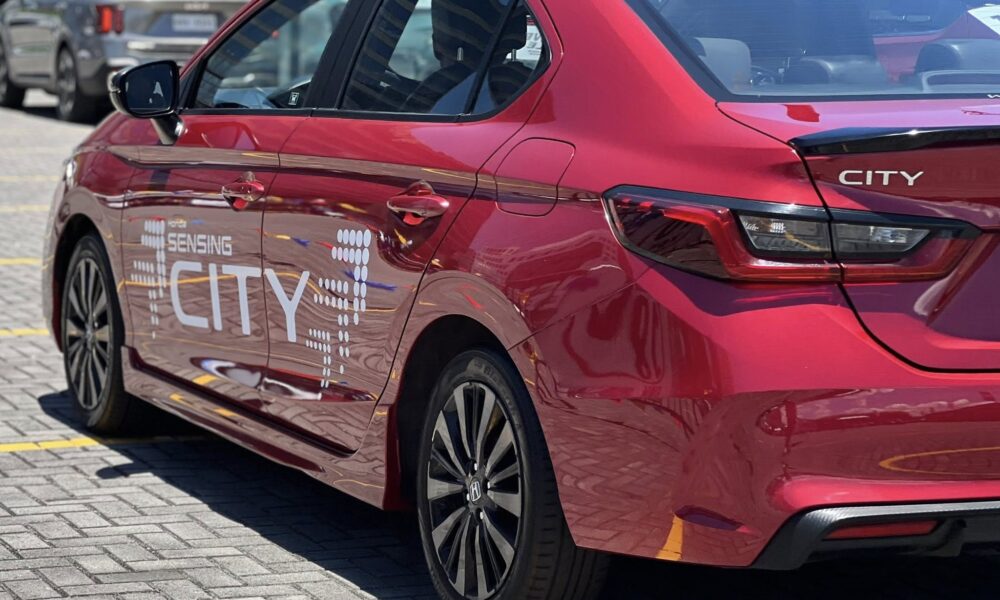 Rear side view of a red Honda City with “Honda Sensing City” graphics parked in a sunlit lot.