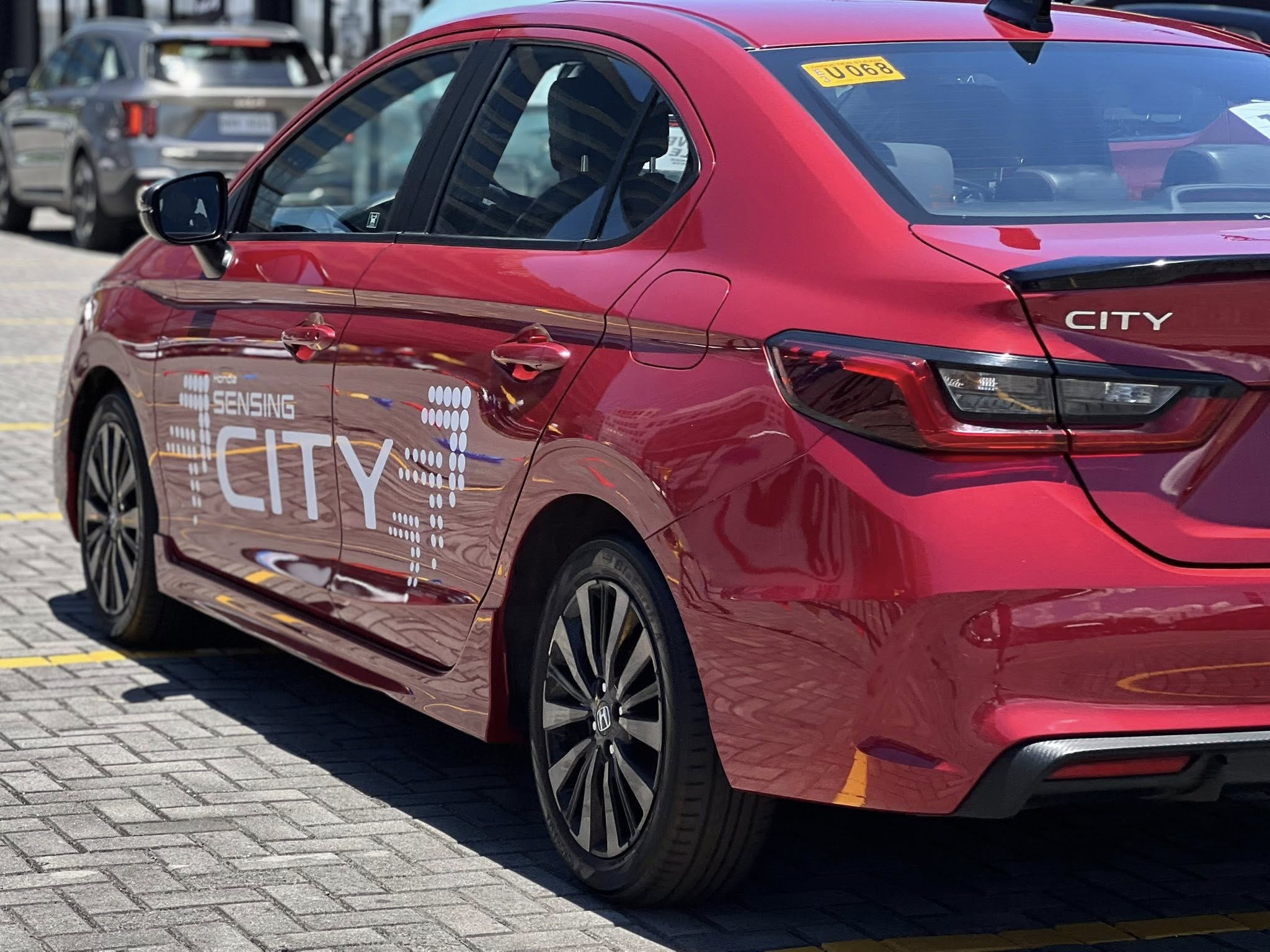 Rear side view of a red Honda City with “Honda Sensing City” graphics parked in a sunlit lot.