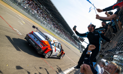 Hyundai N team members cheer and wave flags at the pit wall as the ELANTRA N TCR race car crosses the finish line at the Nürburgring 24-hour endurance race, celebrating a hard-fought victory under bright skies and a packed grandstand.