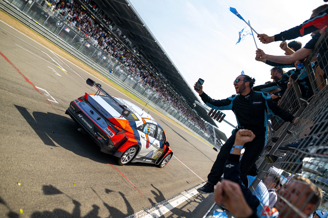 Hyundai N team members cheer and wave flags at the pit wall as the ELANTRA N TCR race car crosses the finish line at the Nürburgring 24-hour endurance race, celebrating a hard-fought victory under bright skies and a packed grandstand.