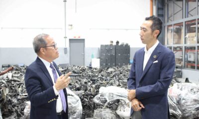 Two men in business attire discuss inside a car dismantling facility with rows of engine parts behind them.