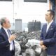 Two men in business attire discuss inside a car dismantling facility with rows of engine parts behind them.