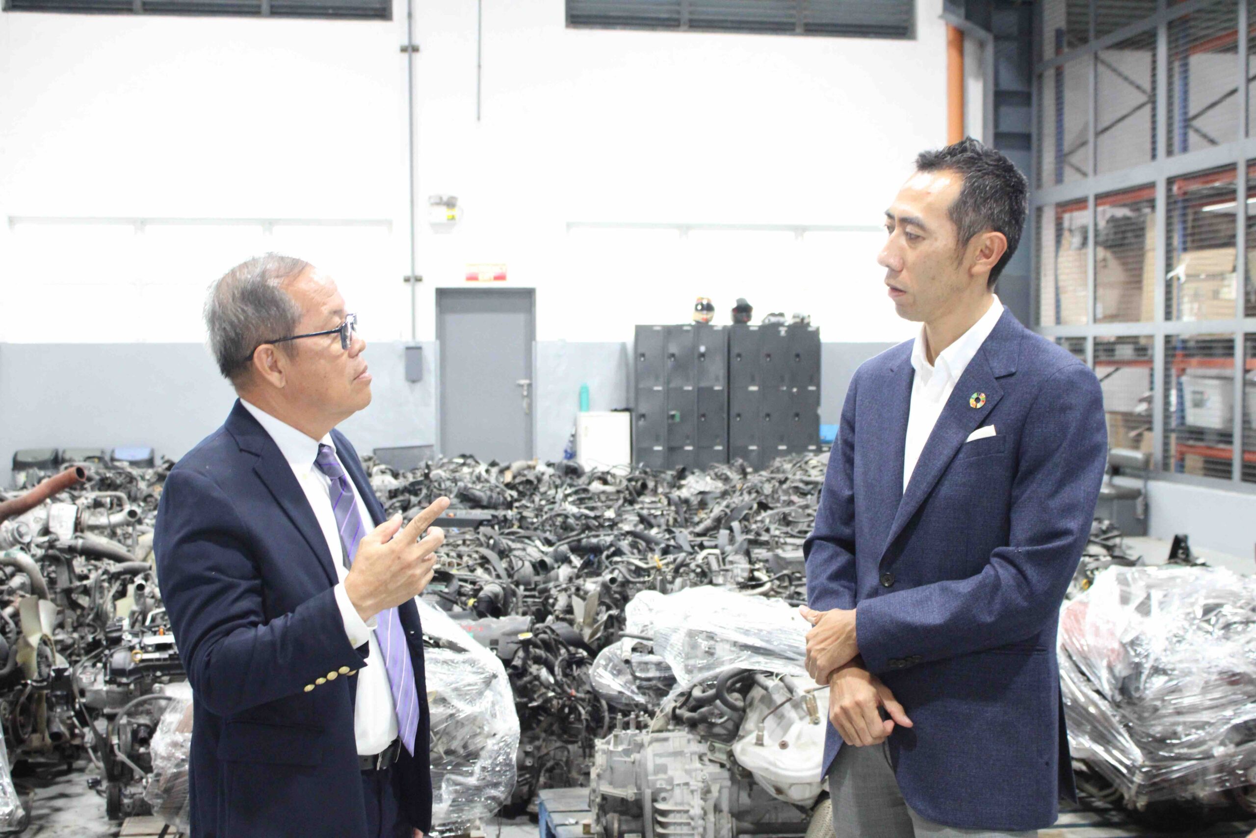 Two men in business attire discuss inside a car dismantling facility with rows of engine parts behind them.