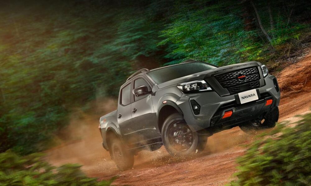 A Nissan Navara PRO-4X drives off-road through a forest trail, kicking up dirt as it climbs a rugged slope.
