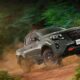 A Nissan Navara PRO-4X drives off-road through a forest trail, kicking up dirt as it climbs a rugged slope.