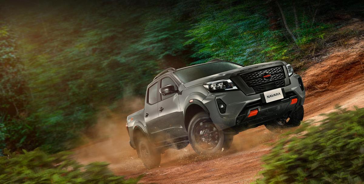 A Nissan Navara PRO-4X drives off-road through a forest trail, kicking up dirt as it climbs a rugged slope.