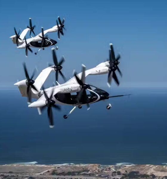 Two Joby electric air taxis in mid-flight over a coastal landscape, demonstrating their vertical takeoff and landing capabilities.