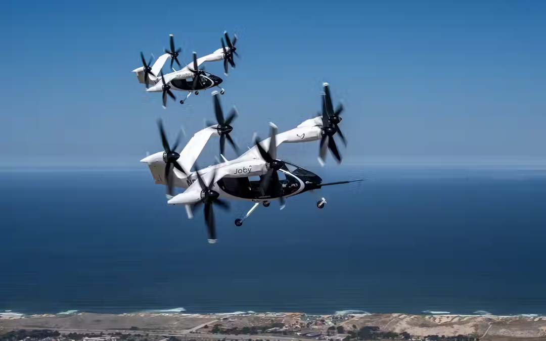 Two Joby electric air taxis in mid-flight over a coastal landscape, demonstrating their vertical takeoff and landing capabilities.