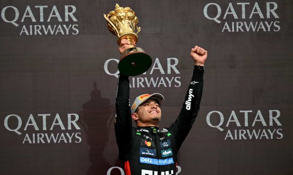 Lando Norris raises a golden trophy and pumps his fist in celebration on the podium after winning the 2025 British Grand Prix, with Qatar Airways branding in the background.