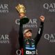 Lando Norris raises a golden trophy and pumps his fist in celebration on the podium after winning the 2025 British Grand Prix, with Qatar Airways branding in the background.