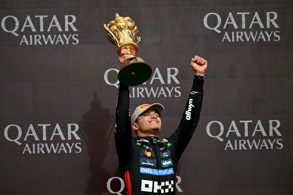 Lando Norris raises a golden trophy and pumps his fist in celebration on the podium after winning the 2025 British Grand Prix, with Qatar Airways branding in the background.