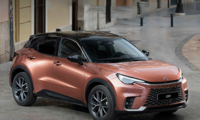 A copper-colored Lexus LBX compact crossover parked on a European-style cobblestone street, featuring sleek headlights, black roof, and sporty alloy wheels.