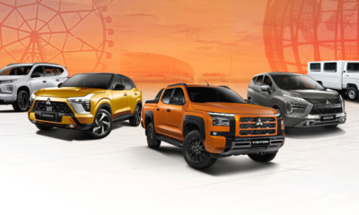 Lineup of Mitsubishi vehicles on display against an orange gradient backdrop featuring a Ferris wheel silhouette. Models shown include the Xpander, Xforce, Triton, Montero Sport, and L300.
