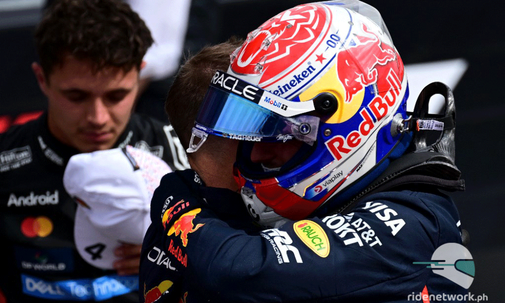 Max Verstappen in full Red Bull racing gear and helmet embraces a team member after qualifying, with McLaren's Lando Norris blurred in the background holding his cap.