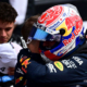 Max Verstappen in full Red Bull racing gear and helmet embraces a team member after qualifying, with McLaren's Lando Norris blurred in the background holding his cap.