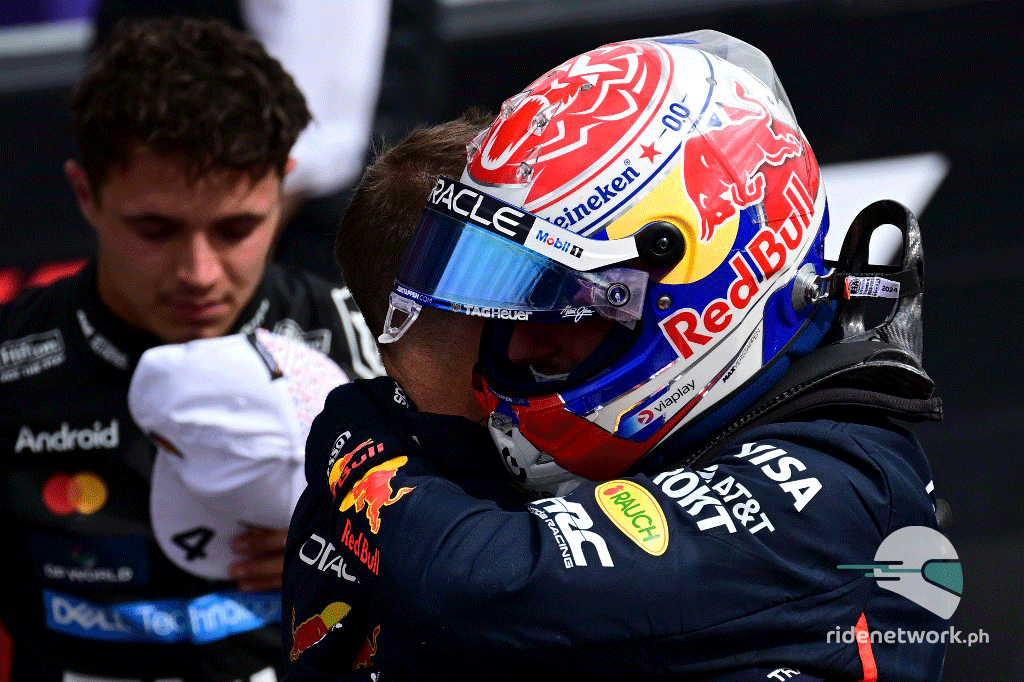 Max Verstappen in full Red Bull racing gear and helmet embraces a team member after qualifying, with McLaren's Lando Norris blurred in the background holding his cap.