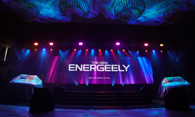 A stage setup with colorful lighting and a large LED screen displaying "The New EnerGeely" with two vehicles under covers on both sides.