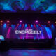 A stage setup with colorful lighting and a large LED screen displaying "The New EnerGeely" with two vehicles under covers on both sides.