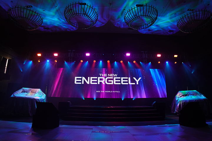 A stage setup with colorful lighting and a large LED screen displaying "The New EnerGeely" with two vehicles under covers on both sides.