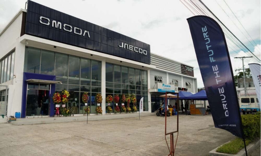 A newly opened Omoda and Jaecoo dealership with a modern glass façade, flower stands at the entrance, and “Drive the Future” banners outside.