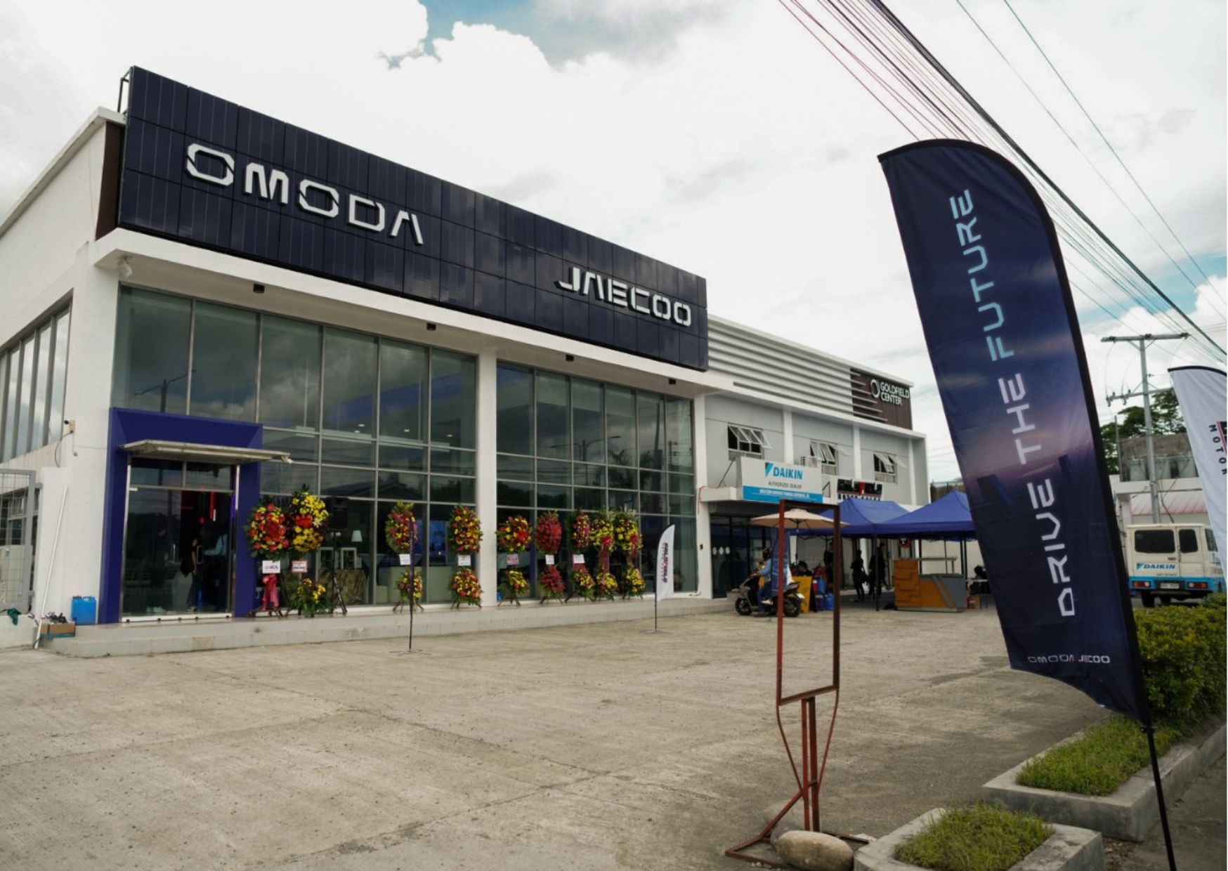 A newly opened Omoda and Jaecoo dealership with a modern glass façade, flower stands at the entrance, and “Drive the Future” banners outside.