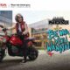 Young man in red varsity jacket sitting on a red Honda NAVi scooter, smiling confidently outside a modern building with “Be Who You WANNAVi” text beside him.