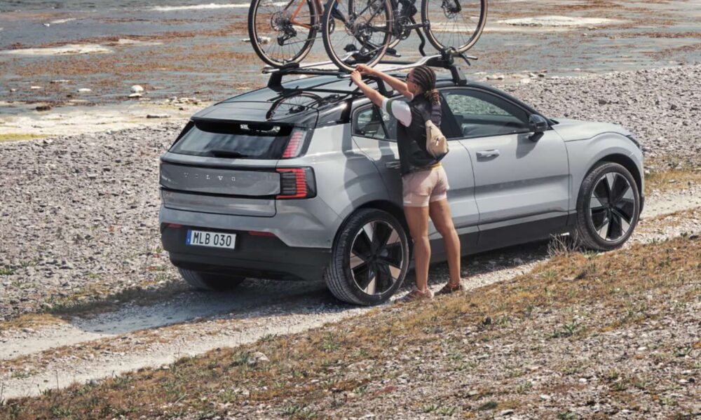 A woman loads a bicycle onto the roof rack of a silver Volvo EX30 parked by a rocky lakeside trail.