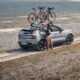 A woman loads a bicycle onto the roof rack of a silver Volvo EX30 parked by a rocky lakeside trail.