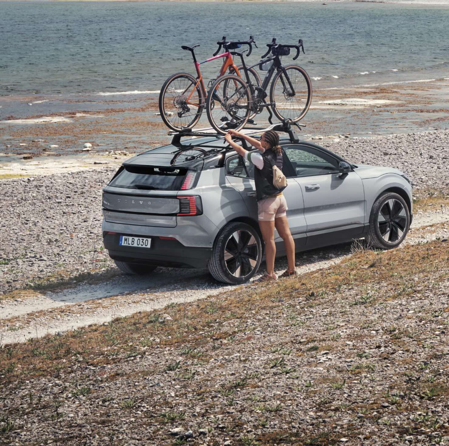 A woman loads a bicycle onto the roof rack of a silver Volvo EX30 parked by a rocky lakeside trail.
