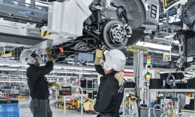 Two automotive assembly workers in safety gear install components underneath a vehicle chassis at a modern car manufacturing facility.