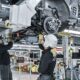 Two automotive assembly workers in safety gear install components underneath a vehicle chassis at a modern car manufacturing facility.
