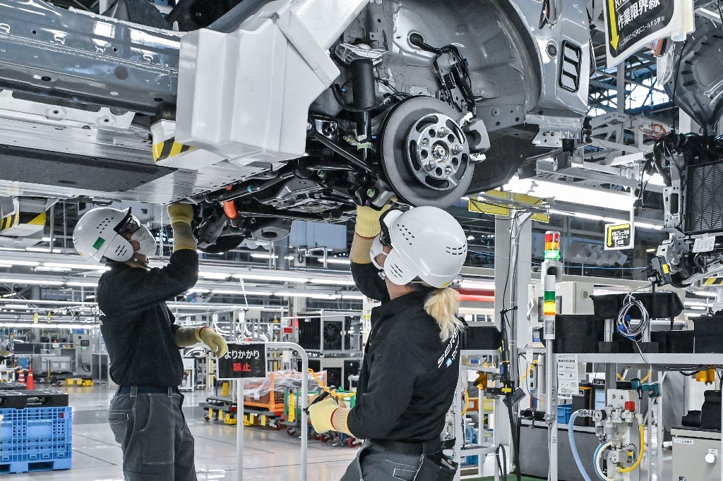 Two automotive assembly workers in safety gear install components underneath a vehicle chassis at a modern car manufacturing facility.
