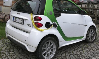 A white and green Smart electric car plugged into a charging station via a yellow cable on a cobblestone driveway.