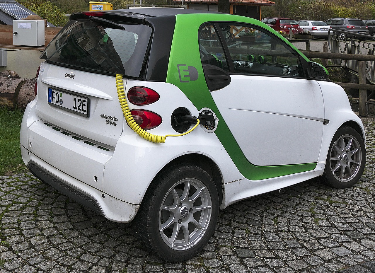 A white and green Smart electric car plugged into a charging station via a yellow cable on a cobblestone driveway.