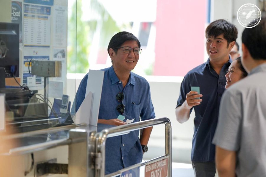 President Ferdinand Marcos Jr. smiles while talking to passengers at a train station booth, holding a fare card, alongside other individuals.