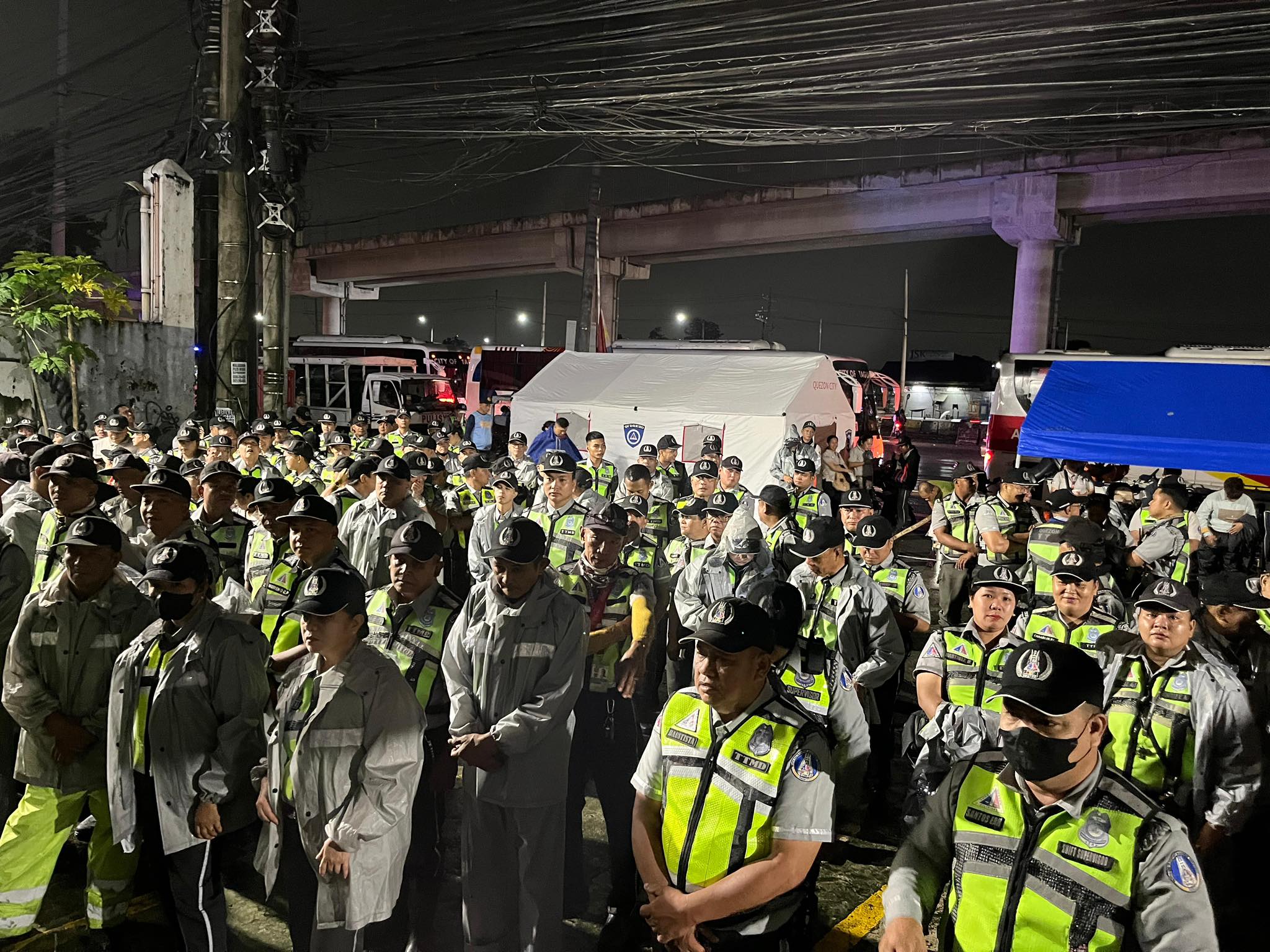 Dozens of uniformed traffic enforcers in reflective vests and rain gear gather at night under bright lights, preparing for deployment ahead of SONA 2025.