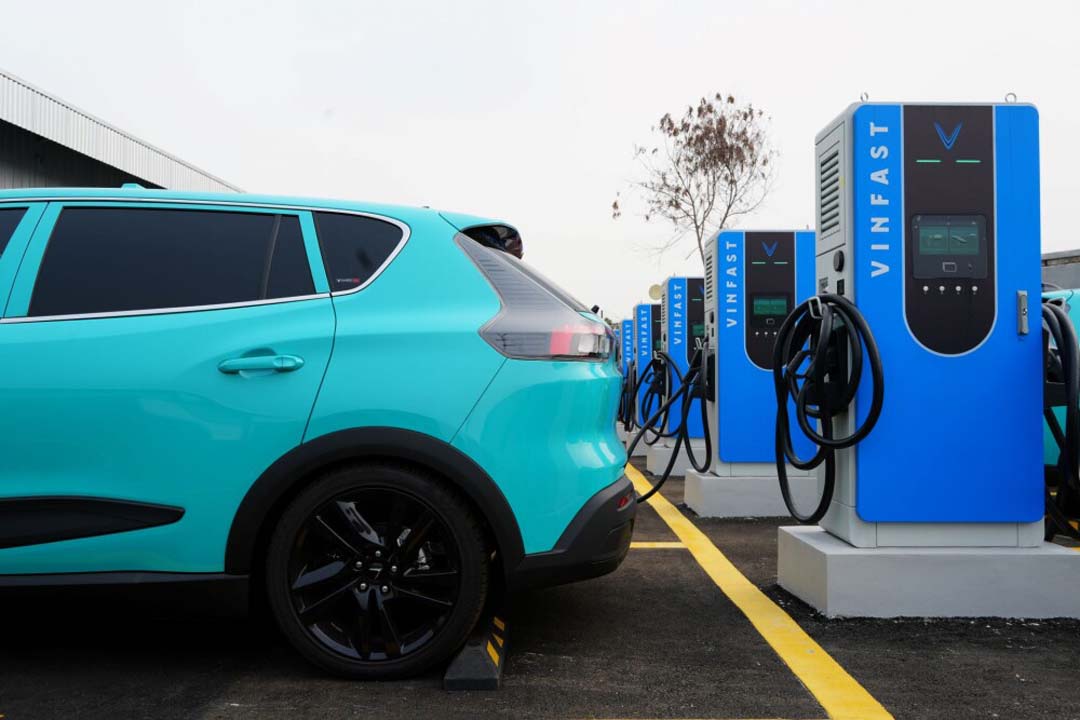 A turquoise VinFast electric vehicle parked and charging at a row of V-Green EV charging stations.